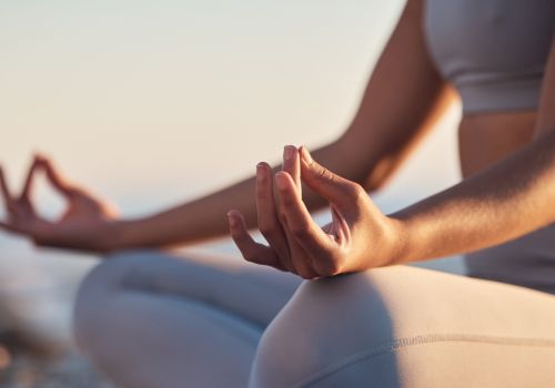 A person in yoga pose outdoors, hands in a mudra/gentle meditation gesture, seated in a calm, sunlit setting by the water.
