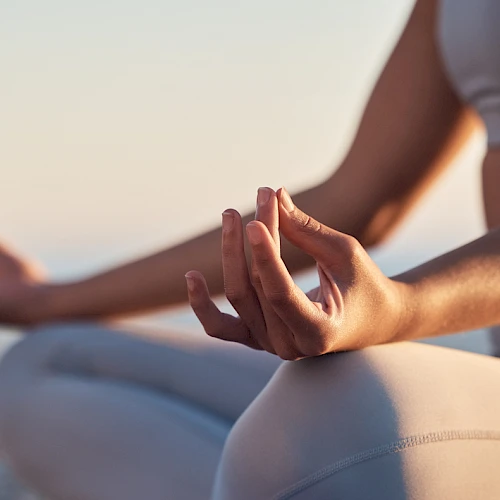 A person in yoga pose outdoors, hands in a mudra/gentle meditation gesture, seated in a calm, sunlit setting by the water.
