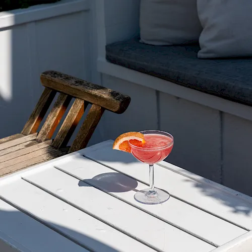 A sunny patio scene with a white bench, a wooden stool, and a pink grapefruit cocktail with a citrus garnish.