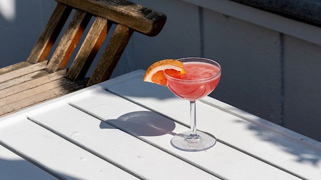 A sunny patio scene with a white bench, a wooden stool, and a pink grapefruit cocktail with a citrus garnish.