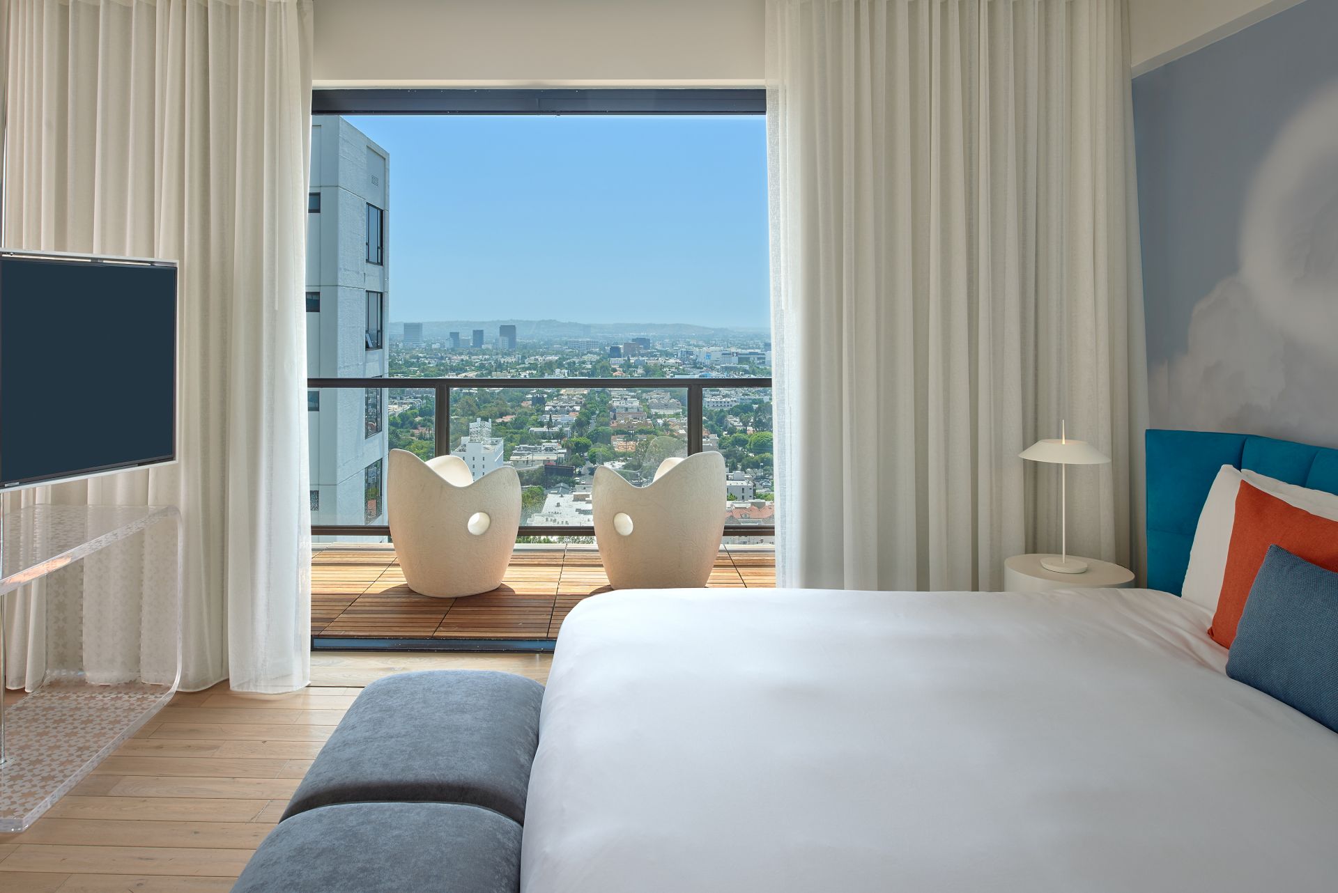 A modern hotel room with a large window balcony, white bed, colorful pillows, a flat-screen TV, and a cityscape view beyond sheer curtains.