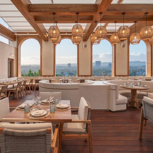 A bright, airy restaurant with wooden beams, warm hanging pendant lights, large windows showing city views, and cozy white and beige seating.