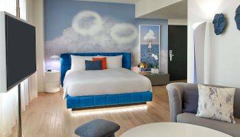 A bright hotel suite with a blue-accent bed, white linens, a circular ottoman, a TV on the wall, and a round table with a notebook, all in a modern, airy space.