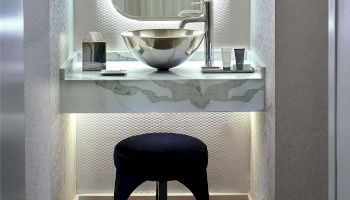 A stylish vanity with a vessel sink, modern faucet, two small containers, a round mirror, and a black cushioned stool under soft lighting.