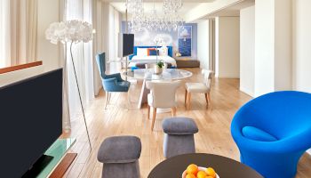 A bright, modern living room with a round table and bowl of oranges in the foreground, blue chair, gray stools, and a chandelier overhead.