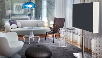 A futuristic living room with a large flat-screen TV, white modern chairs, a black pouf, glass desk, and a blue-tinted, tunnel-like art installation in the back.
