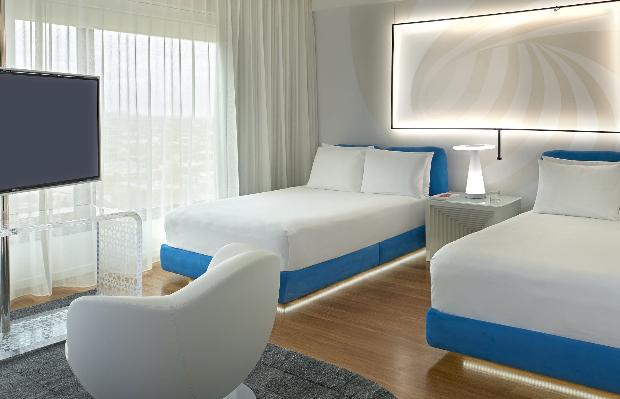 A bright hotel room with two white beds, blue accents, a chair, a flat-screen TV, and large windows letting in natural light.