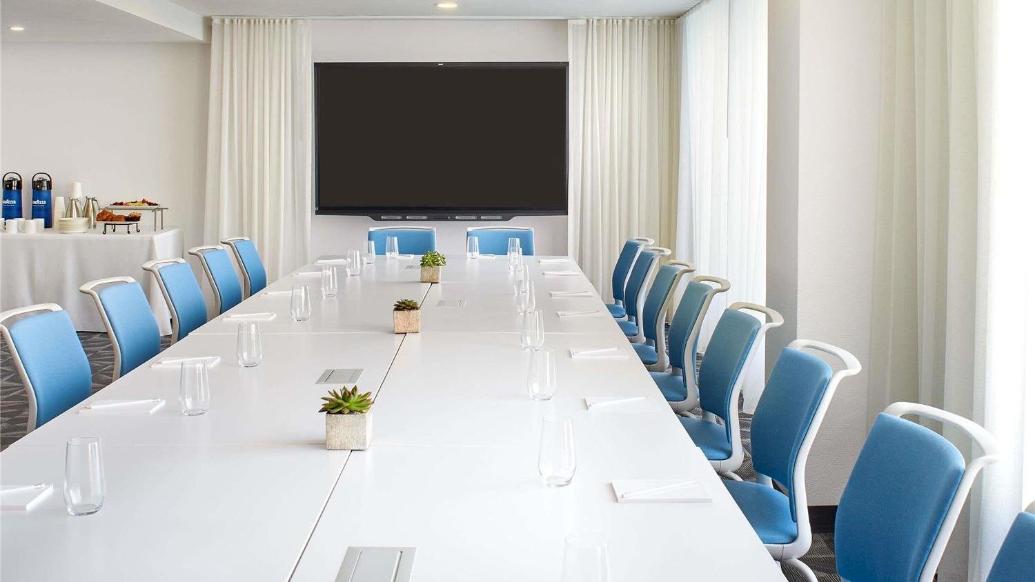 A modern conference room with a long white table, blue chairs, a large screen at the far end, and natural light from windows on both sides.