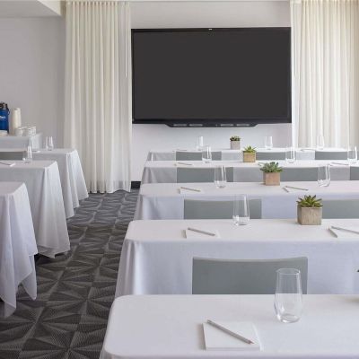 Cozy conference room set for a seminar: rows of white-clothed tables with notepads, pens, glasses, and small plants, a large screen at the front.