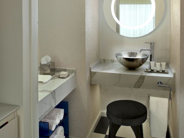A compact, modern bathroom with a round backlit mirror, vessel sink, black stool, checkered floor, and a small vanity counter with neatly folded towels.