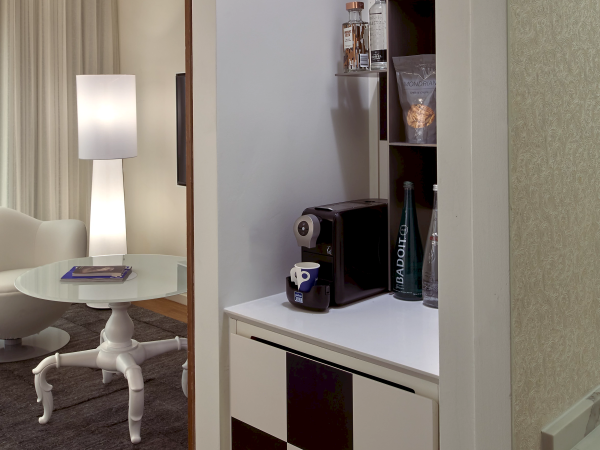 A compact home bar/coffee station setup with a built‑in cabinet, a small coffee machine, glassware, a mug, and a checkerboard cabinet door, in a cozy living nook.