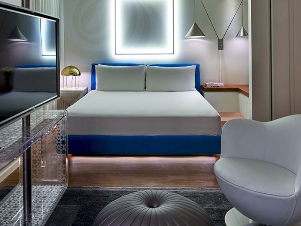 Luxurious hotel room with a bright square mirror light above a white bed, blue accents, modern seating, and a sleek, contemporary vibe.