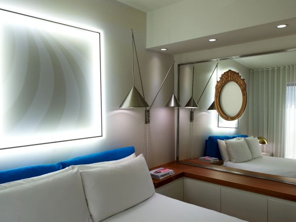A modern hotel room with a large backlit square art piece, mirrored wall, pendant lights, a blue bed runner, white pillows, and a round ornate mirror.