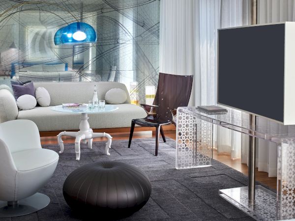 A modern living room with white furniture, a large wall-mounted TV, a black pouf, and a bright blue arcade-like light fixture in a stylish, contemporary space.