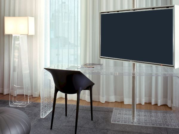 A modern living room setup with a transparent acrylic desk and TV stand, a black chair, a wall-mounted flat screen, carpet, and sheer curtains.