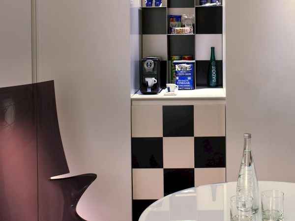 A modern lounge area with a black-and-white checkerboard feature wall and shelf display, a purple chair, and a white round table with glass bottles, at 140 characters.