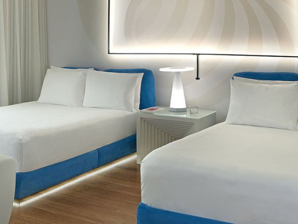 Two modern hotel beds with white linens, blue bases, a nightstand, lamp, and a soft-lit wall art panel above.