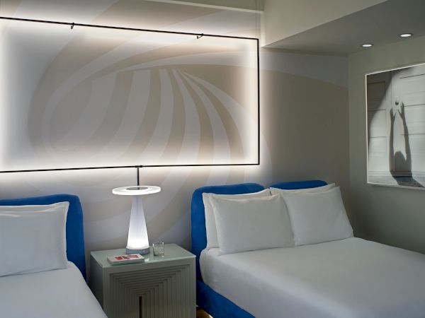Two beds with white linens and blue headboards in a modern hotel room, a nightstand with a lamp between them, framed art on the wall, and a backlit framed ceiling pattern.