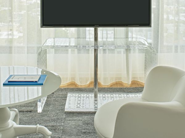 A modern office lounge with a mounted flat-screen TV on a stand, curved white chair, small table, and light sheer curtains in the background.