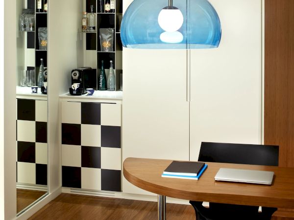 A stylish home office with a round wooden table, black chairs, a blue dome pendant light, and a checkerboard cabinet reflected in a mirrored wall.