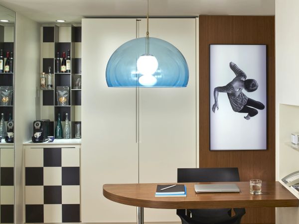A modern home office with a curved wooden desk, a blue pendant lamp, a black-and-white checkered cabinet to the left, and a framed black-and-white photo on the wall.
