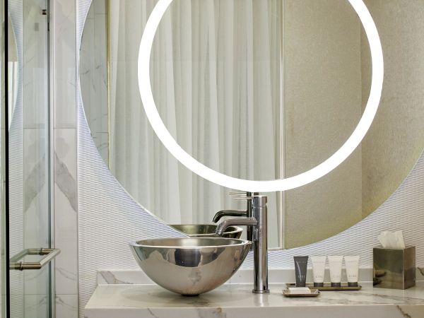 A modern bathroom with a circular backlit mirror above a marble sink, glass shower to the left, and neatly stacked towels beneath the counter.
