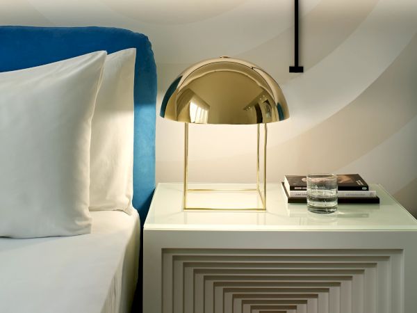 A stylish bedroom nightstand with a geometric stack of nested white drawers, a brass dome lamp, a bed with white linens, and a striped wall pattern.
