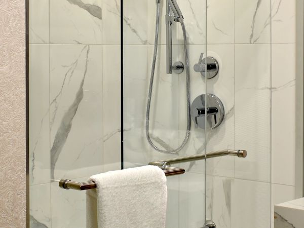 A modern glass shower enclosure with a handheld shower, mounted rail, and a towel bar across the glass; white tile walls behind.