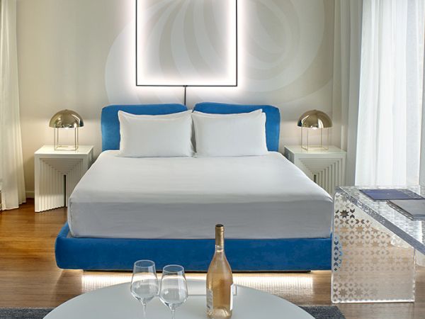 A modern hotel room with a plush blue and white bed, round white table set with a bottle and glasses, and soft lighting in a bright, airy space.