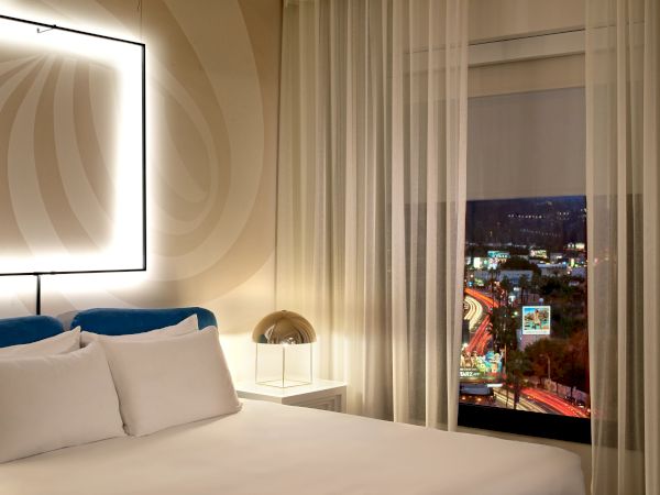 A stylish hotel room with a large bed, white linens, blue pillows, soft lighting, sheer curtains, and a city view at night.