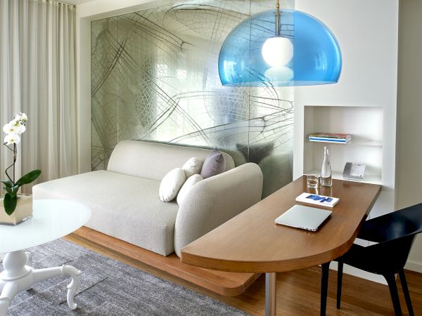A cozy hotel-like living area with a curved desk, a light gray sofa, a blue pendant light, a white coffee table, and a small shelf unit with a lamp.