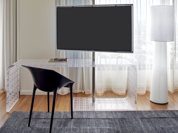 A modern home office setup with a wall-mounted TV, a glass desk, a black chair, sheer curtains, a tall floor lamp, and a dark area rug.
