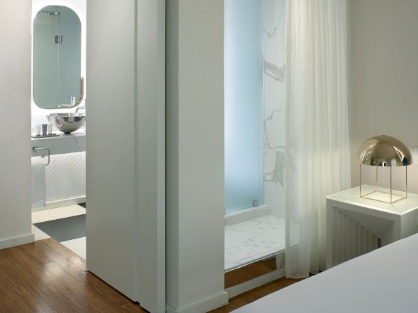 A bright hotel-style bedroom with an open en-suite bath and separate shower, featuring white walls, a wooden floor, and soft lighting.