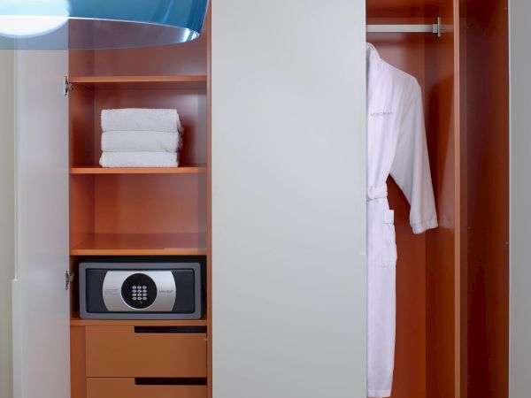 A modern open wardrobe split by a white panel: shelves with towels, a small safe, drawers, and a hanging space for clothes, all beside a curved blue light.