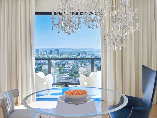 A bright, modern dining nook with a round glass table, crystal chandeliers, white drapes, blue sky view, and stylish white and blue chairs.