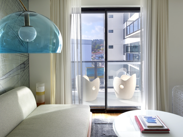 A modern living room with a light sofa, a large blue pendant lamp, a glass door leading to a balcony with two chairs, and a round coffee table with magazines, ending with a cityscape view.