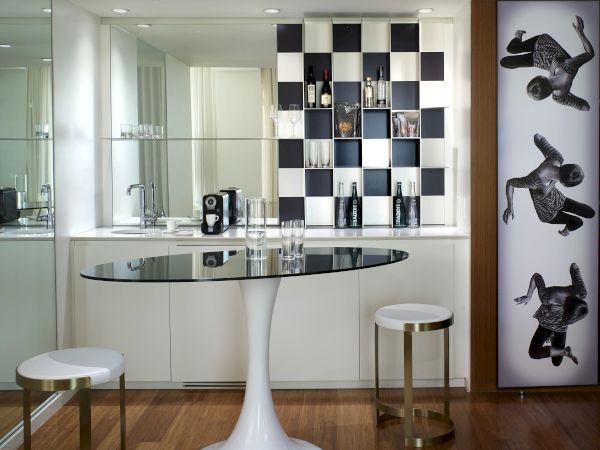 A modern kitchen lounge with a round glass table, white stools, a black-and-white checkered wall display, and art panels of riders on the right.