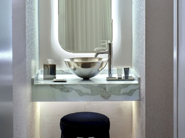 A chic dressing area with a backlit arched mirror, marble countertop, metallic sink, and a dark-stained stool under soft ambient light.