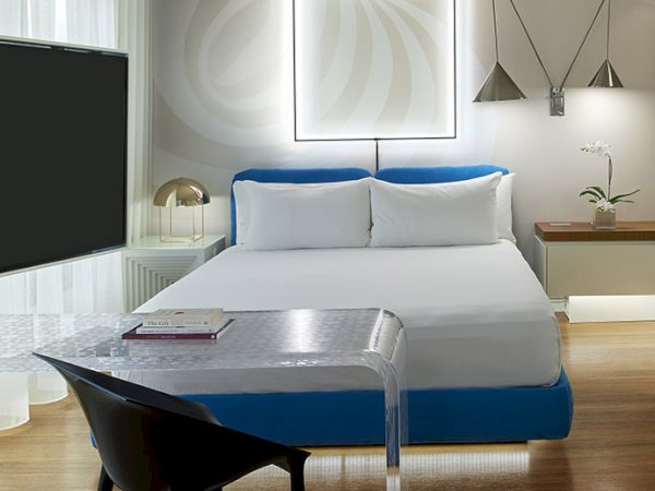 Modern hotel room with a king bed, blue accents, glass desk, black chair, wall art with neon square, warm wood floor, couch and desk lamp.