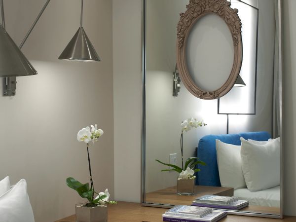 A modern bedroom corner with a built-in wooden bench, white cushions, hanging pendant lights, a large mirror, an ornate oval frame on the wall, and potted orchids on the surface.