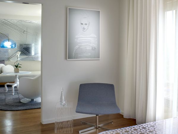 A modern, minimalist living area with a gray lounge chair, framed portrait, sheer curtains, and a bright adjacent room featuring a blue pendant light.