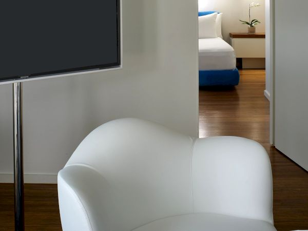 A modern hotel room with a white curved chair, a round white table with an orange book, and a doorway to a neatly made bed in the background.