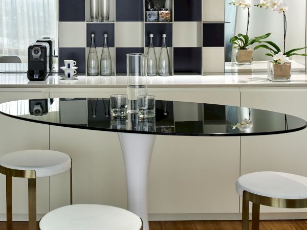 A modern kitchen nook with a checkered wall, backlit shelves, a round glass table, white stools, and a sleek white pedestal table.