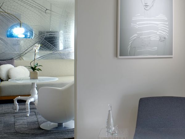 A modern, minimalist living space with a white seating area, a gray chair, and a framed monochrome portrait on the wall, plus a small glass table and decorative sculpture.