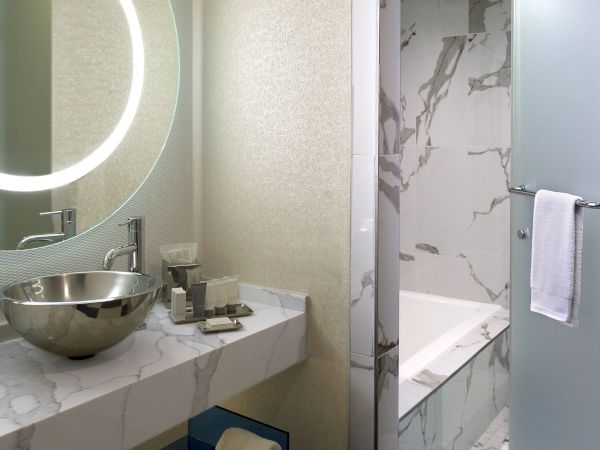 A modern bathroom with a round LED mirror above a marble countertop, a vessel sink, and a glass-enclosed shower with marble tiles and a towel rack.