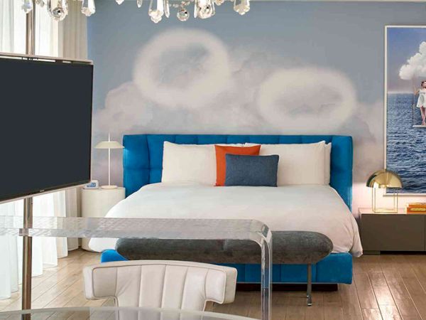 A stylish hotel room with a blue upholstered bed, fluffy white bedding, an orange and gray accent pillow, chandelier, chandelier, wall art, and a bowl of oranges on a glass table.