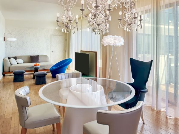 A modern, luxurious living area with a round glass dining table, stylish chairs, chandeliers, and a bright, airy lounge behind, featuring a sofa and a TV.