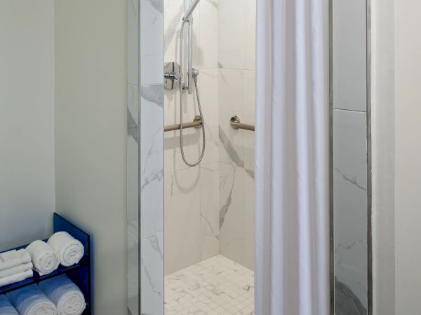 Shower stall with a white curtain, marble walls, a handheld shower, and a small window, adjacent to a towel rack with rolled towels.