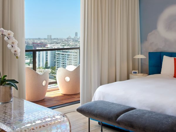 A chic hotel room with a view: white bed, blue accent wall, bedside lamps, and a balcony with white chairs overlooking a city skyline, bright and serene.