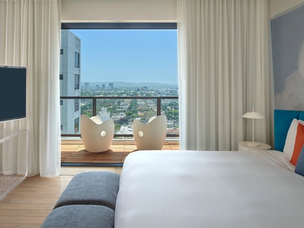 A modern hotel room with a large bed, white linens, colorful pillows, a TV, and a balcony with two white chairs overlooking a cityscape.